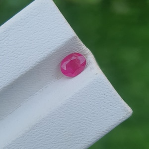 May include: A faceted, oval-shaped ruby gemstone. The ruby is a vibrant, deep pinkish-red color. It rests on a textured, white surface, with a blurred green background.