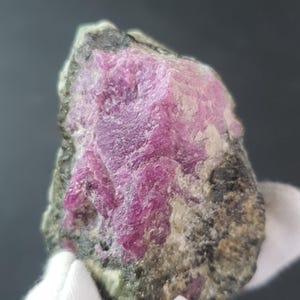 Pode incluir: Um espécime mineral bruto com uma vibrante estrutura cristalina rosa intenso. A área rosa é circundada por uma matriz de tons cinza, preto e verde. A pedra natural tem uma superfície áspera e texturizada, mostrando seu estado bruto.