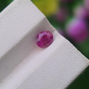 May include: A faceted, oval-shaped ruby gemstone. The ruby is a deep, vibrant red color and is set against a textured white surface. The background is blurred, with hints of green and purple.