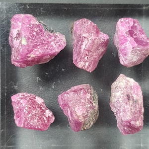 May include: Six rough-cut, raw ruby gemstones. The rubies are a vibrant pinkish-red color with a crystalline texture. Each stone has a unique shape and size, with some showing dark gray inclusions. The rubies are displayed on a black surface.