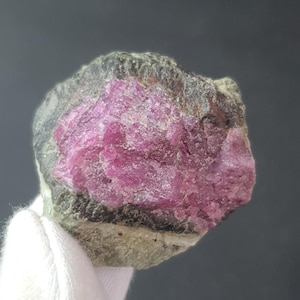 Puede incluir: Una gema en bruto, sin cortar, con un interior rosa vibrante y un exterior oscuro y texturizado. La piedra es sostenida por una mano enguantada de blanco, mostrando su belleza natural y en bruto. El color de la gema contrasta con el fondo oscuro.