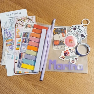 Könnte beinhalten: Eine Sammlung von Schreibwaren für Bücherliebhaber, darunter ein Lesezeichen, Haftnotizen, Stifte, Aufkleber, Washi-Tape und ein Bleistift. Die Artikel sind in Lila, Orange und Weiß gehalten, mit dem Namen "Marina" in lila Buchstaben.