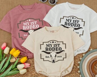 May include: Three baby bodysuits in dusty rose, cream, and white, each featuring a "My 1st Rodeo" graphic with a bull skull and the year 2024. The bodysuits are short-sleeved and have a round neckline. Sunglasses and flowers are in the background.