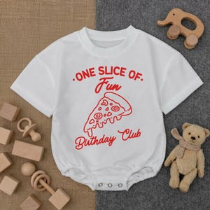 May include: White baby bodysuit with a red pizza slice graphic and the text "ONE SLICE OF. Fun Birthday Club". The short-sleeved onesie has a round neckline and snap closures at the bottom. Wooden toys and a teddy bear are in the background.