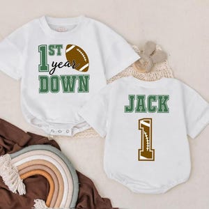 May include: Two white baby onesies with football-themed designs. One features the text "1st year DOWN" with a football graphic. The other has the name "JACK" and the number "1" with a football design.