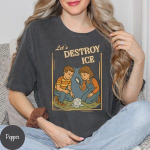 May include: Dark gray t-shirt with the text "Let's DESTROY ICE" above an illustration of two children examining a melting ice cube with a magnifying glass. The children are wearing orange and blue clothing.