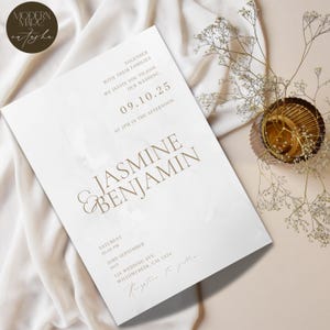 May include: A wedding invitation featuring the names Jasmine & Benjamin in gold lettering. The invitation includes the date, time, and location details, set against a white background. The invitation is placed on a cream-colored fabric.