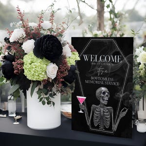 May include: A black sign with a coffin-shaped frame that reads "WELCOME, THIS IS A FUNERAL, it's a BOTTOMLESS MEMORIAL SERVICE." A skeleton holds a pink cocktail. A white vase holds a bouquet of black, white, and green flowers.