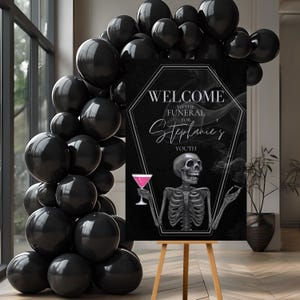May include: A black sign on a wooden easel with the text "WELCOME TO THE FUNERAL FOR Stephanie's YOUTH". The sign features a skeleton holding a pink cocktail. Black balloons are arranged around the sign.
