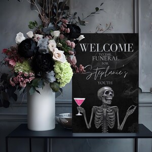 May include: A black sign with the text "WELCOME TO THE FUNERAL FOR Stephanie's YOUTH" and a skeleton illustration holding a pink cocktail. A white vase with a floral arrangement of black, white, and pink flowers sits to the left.