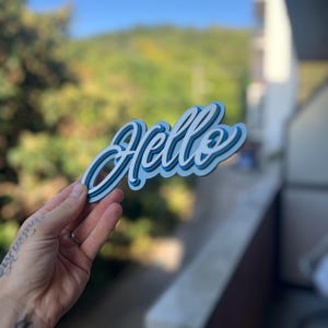 May include: A layered, three-dimensional sign featuring the word Hello in a cursive script. The design consists of stacked layers in shades of light blue, medium blue, and dark blue, creating a depth effect with a clean, bold outline.