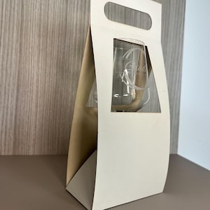 Op de afbeelding: Een witte kartonnen wijnglas geschenkdoos met een rechthoekig venster met een helder wijnglas. Op het wijnglas staat het woord "Grad" geëtst. De doos heeft een handvat aan de bovenkant en staat op een bruin oppervlak.