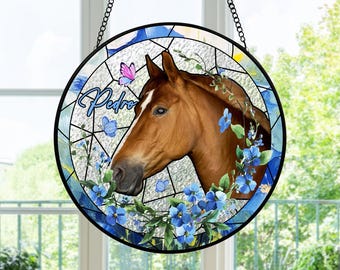 Pode incluir: Janela redonda de vitral com a cabeça de um cavalo castanho com uma risca branca, rodeada por flores e borboletas azuis. O nome "Pedro" está escrito em itálico. A janela é emoldurada em preto e pendurada por uma corrente.