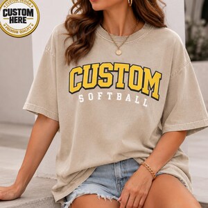 May include: A beige short-sleeved t-shirt with the word "CUSTOM" in yellow block letters with a black outline, above the word "SOFTBALL" in white. The shirt has a vintage look and a "CUSTOM DESIGN" logo in the upper left corner.