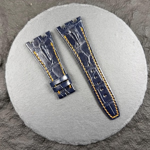 May include: Two navy blue watch straps with yellow stitching. The straps are made of a textured material, possibly leather, and are designed to attach to a watch face. The straps are displayed on a grey, circular surface.