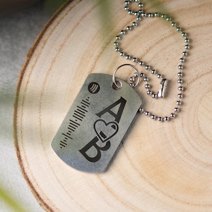 Könnte beinhalten: Silberner Dog-Tag-Anhänger mit einer Kugelkette. Der Anhänger zeigt einen Spotify-Code, die Buchstaben "A" und "B" mit einem Herzsymbol und eine Reihe vertikaler Linien. Der Anhänger wird auf einer Holzoberfläche präsentiert.