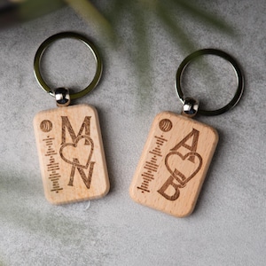 Könnte beinhalten: Zwei Holzschlüsselanhänger mit silbernen Schlüsselringen. Jeder Schlüsselanhänger hat eine rechteckige Form mit eingravierten Initialen und einem Herzsymbol. Einer hat die Buchstaben "M" und "N", der andere "A" und "B". Beide haben ein Schallwellendesign.