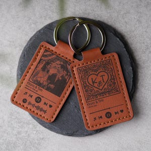 Könnte beinhalten: Zwei braune Leder-Schlüsselanhänger mit silbernen Ringen. Einer zeigt ein Foto und den Text "Klara & Tim love in the air" mit Musiksymbolen. Der andere hat ein Herzdesign mit den Initialen "K&T" und dem Text "Klara & Tim I love you".