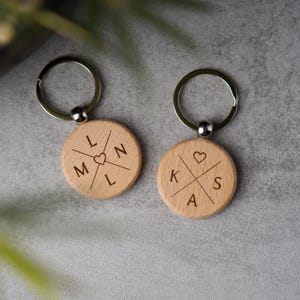 Könnte beinhalten: Zwei Holzschlüsselanhänger mit silbernen Ringen. Jeder Schlüsselanhänger hat einen runden, hellfarbigen Holzanhänger mit eingravierten Initialen und einem Herzsymbol. Die Initialen sind kreuzförmig angeordnet.