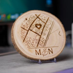 Könnte beinhalten: Eine runde Holzscheibe mit einer gelaserten Stadtkarten-Gravur, einem Herzsymbol und den Initialen "M&N". Das Holz hat eine natürliche, hellbraune Farbe und wird auf einem transparenten Ständer präsentiert. Die Karte zeigt Straßen und Gebäude.