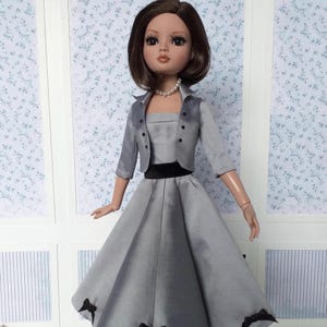 May include: A doll in a grey dress with a matching jacket and a black belt. The dress features a full skirt with black bows at the hem. The doll has brown hair and a pearl necklace.