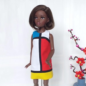 May include: A doll in a sleeveless dress with a colour block design. The dress has a white central panel with a black vertical stripe and a black horizontal stripe at the top. The dress has a blue top, red side panel, and yellow hem.