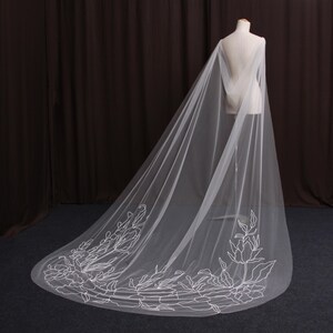 Peut inclure: Un long voile de mariée blanc et fluide avec une broderie florale sur le bord inférieur. Le voile est drapé sur un mannequin, mettant en valeur sa longueur et son design délicat. La broderie présente des motifs floraux et de feuilles blancs détaillés.