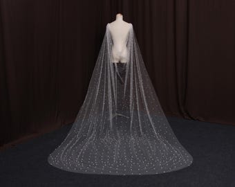 May include: A sheer, white bridal cape with a multitude of small, evenly spaced pearl-like embellishments. The cape is draped over a mannequin, showcasing its flowing design and long train. The background is a dark brown curtain.