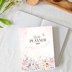 Peut inclure: Un agenda quotidien 2026 sur le thème floral avec un design à l'aquarelle. La couverture présente le texte "My daily PLANNER" et une bordure florale avec des fleurs roses et violettes. L'agenda est ouvert sur une page blanche.