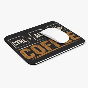 May include: A rectangular black mouse pad featuring the white text CTRL + ALT and a graphic of a coffee cup, with the large brown text COFFEE printed below. A smooth white computer mouse rests on the surface of the mouse pad.