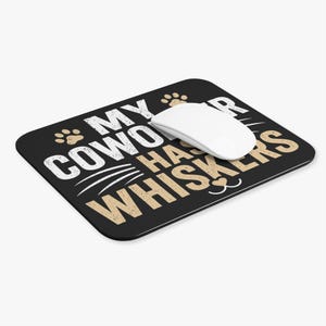 Peut inclure: Un tapis de souris rectangulaire noir avec le texte blanc et beige MY COWORKER HAS WHISKERS, orné d'empreintes de pattes et d'un graphique de museau de chat, avec une souris d'ordinateur blanche posée sur la surface du tapis.