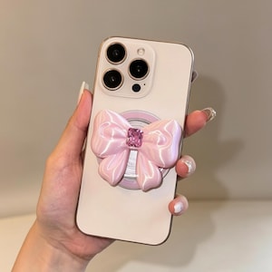 Pode incluir: Um smartphone de cor clara com um suporte circular na parte traseira. O suporte é adornado com um laço tridimensional rosa claro brilhante, centrado com uma única pedra preciosa de cristal rosa retangular e facetada.