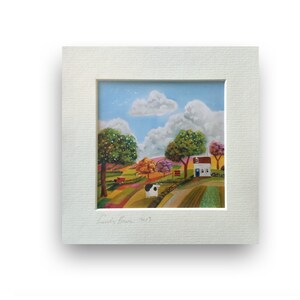 May include: A framed painting depicting a colourful countryside scene with a white house, green trees, a red wagon, and a black and white cow. The painting is signed "Lucy's Barn 2017".