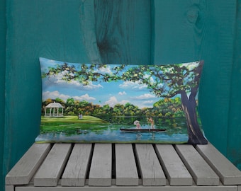 May include: A decorative pillow featuring a painting of a lake scene with a couple in a rowboat, a gazebo in the background, and lush green trees. The sky is blue with white clouds.