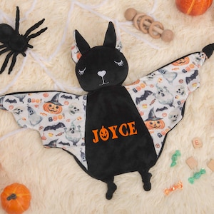 Puede incluir: Una manta para bebé con forma de murciélago negro con el nombre "JOYCE" en naranja. Las alas presentan un estampado de Halloween con calabazas, fantasmas y murciélagos. Una araña negra y calabazas están cerca.