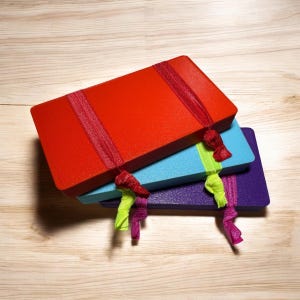 May include: Three rectangular notebooks stacked on top of each other. The top notebook is red, the middle is blue, and the bottom is purple. Each notebook has a ribbon closure with a decorative knot. The background is a light wood grain.