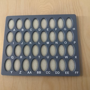 May include: A dark gray alphabet display board with 30 oval openings, each containing a beige insert. The letters A through FF are printed above or below each opening. The board is rectangular and sits on a light brown wooden surface.