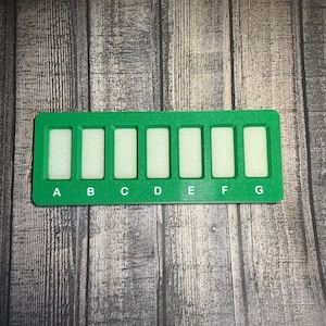 May include: A rectangular green plastic organizer tray featuring seven vertical slots, each containing a light-colored foam insert. Below each slot, the letters A, B, C, D, E, F, and G are printed in white, providing a clear labeling system.