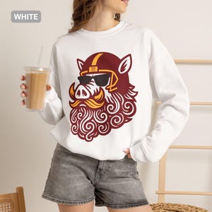 May include: A white long-sleeve sweatshirt featuring a graphic of a stylised boar wearing a football helmet and sunglasses. The boar has a prominent curly beard and moustache in shades of maroon and gold. The text WHITE is visible in the corner.