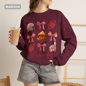 May include: A maroon long-sleeve sweatshirt featuring a graphic print of a hog wearing a beanie and sunglasses, surrounded by footballs, bows, and a foam finger. The text Washington is printed in a script font at the bottom of the design.