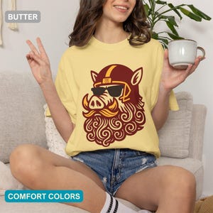 May include: A butter yellow t-shirt featuring a graphic of a wild boar wearing a maroon and gold football helmet and black sunglasses. The boar has a stylised curly beard and moustache. The text BUTTER and COMFORT COLORS appear in labels.