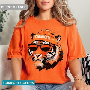 May include: A burnt orange t-shirt featuring a graphic of a tiger wearing a matching orange beanie with the word CINCINNATI printed in white and black sunglasses. The tiger has black stripes and white whiskers on a vibrant orange background.