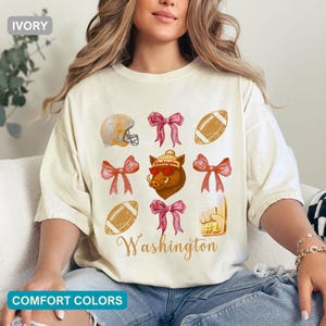 May include: An ivory t-shirt featuring a grid of graphics including a football helmet, pink bows, footballs, a foam finger with the number 1, and a boar wearing a beanie and sunglasses, with the word Washington printed in cursive script.
