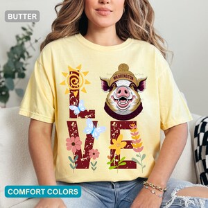 Pode incluir: Uma t-shirt amarelo-manteiga com um gráfico da palavra LOVE em vermelho escuro, onde o O é substituído por um porco com um gorro bege com a inscrição WASHINGTON. O design inclui um sol amarelo, borboletas azul-claras e flores coloridas.