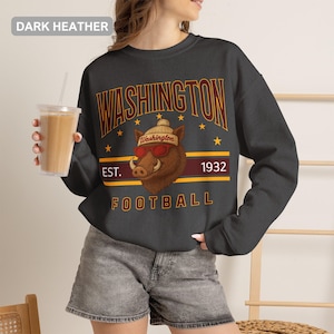 Pode incluir: Uma camisola de gola redonda em cinzento escuro mesclado com um gráfico de um javali castanho a usar um gorro e óculos de sol vermelhos. O texto diz WASHINGTON, EST. 1932 e FOOTBALL em letras amarelas e vermelhas, com pequenas estrelas.