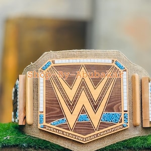 Könnte beinhalten: Ein hölzerner WWE World Heavyweight Championship-Gürtel mit braunem und goldenem Design. Der Gürtel zeigt das WWE-Logo, türkisfarbene Akzente und den Text "WORLD HEAVYWEIGHT CHAMPION". Der Text "Shop By - Abubakar" ist ebenfalls sichtbar.