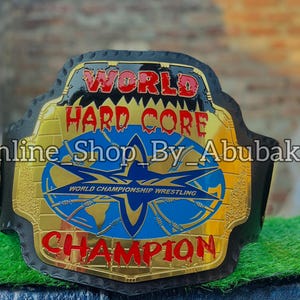 Puede incluir: Un cinturón de campeonato de lucha libre negro y dorado con las palabras "WORLD HARD CORE CHAMPION" en letras rojas y doradas. El cinturón tiene un emblema azul y dorado con las palabras "WORLD CHAMPIONSHIP WRESTLING". También se ve el texto "Online Shop By _Abubakar".