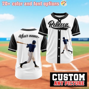 May include: White baseball jersey with black sleeves and collar. The front features the name "Rebecca" in black script, and a baseball player graphic. The back has "Your name" in script and a baseball player graphic. Text at the top says "70+ color and font options".