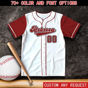 Puede incluir: Una camiseta de béisbol blanca con mangas granates, con el nombre "Rebecca" en fuente cursiva y el número "00". La camiseta tiene ribetes rojos y es abotonada. También se ven una pelota de béisbol, un bate y el texto "70+ COLOR AND FONT OPTIONS" y "CUSTOM ANY REQUEST".
