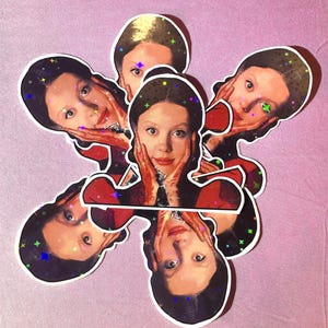 Può includere: Una collezione di adesivi raffiguranti il ritratto di una persona con capelli scuri e un abito rosso, con le mani sul viso. Gli adesivi hanno un effetto olografico e glitterato e sono disposti su uno sfondo rosa.